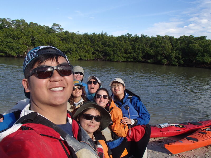 A group of people are posing for a picture in front of a body of water. They appear to be on a kayaking trip, as some kayaks are visible in the background. The people are wearing casual clothing and some have hats and sunglasses. The background features lush green trees and a calm body of water, suggesting a natural setting. The overall atmosphere seems cheerful and adventurous.